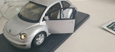 The image displays a silver Volkswagen New Beetle model car, positioned on a black rectangular display base. The model is angled, presenting its front and left side, with the driver's side door open, revealing a light-colored (possibly beige or light gray) interior. Notable details on the car include the prominent circular Volkswagen (VW) logo on the front hood, circular headlights with a yellowish hue, and multi-spoke silver wheels. The surface of the model car shows some visible dust accumulation.

The scene appears to be indoors, likely on a plain white surface, such as a table or shelf. In the blurry background to the left, a translucent glass object is partially visible. To the right, a white and green card or paper object is partially visible; it contains some indistinct, blurry text with discernible letters like "ge" and a small, blurry illustration. There are no people or activities taking place; the image is a static shot of the car model. No specific time of day or weather can be inferred. The location context provided for the media file is Beringen, Belgium, though no explicit visual cues within the image confirm this.