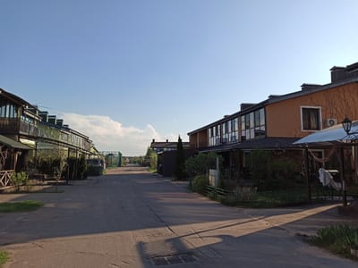 The image depicts a residential street in Рыбхоз, Russia. The street is paved and stretches into the distance, flanked by two-story buildings on either side. The buildings on the left are clad in green siding and feature balconies. The buildings on the right are of brick construction, with large windows. Both sides have mature trees and shrubbery. The sky is a clear blue with some clouds visible in the distance. The light suggests it is daytime, with shadows cast across the street. The scene appears quiet and residential.