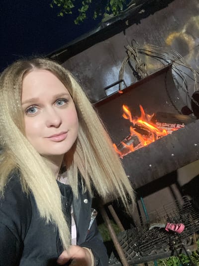 A selfie-style image captures a young woman with long, light blonde, crimped hair and blue eyes, looking directly at the camera. She has light makeup, including pink lipstick and eye makeup, and is wearing a dark jacket over a lighter, possibly plaid, garment. Her left hand is partially visible near her shoulder.

The scene appears to be outdoors or in a dimly lit, semi-enclosed outdoor space, likely at night or during late evening, given the darkness and reliance on the fire for illumination. Behind the woman, and slightly to her upper left, is a metal fire pit or burner containing bright orange flames and burning wood. To the left of the fire, a tangled bundle of thick wires or cables rests on a dark ground surface, which shows some yellow staining. To the right of the fire, a black metal grill or grate structure is visible, with a pink-handled knife or tool inserted into it. Bits of green foliage can be seen in the bottom left and top right corners of the background.

The activity depicted is a casual moment, possibly a selfie taken during an outdoor gathering or a relaxed evening by a fire. The woman’s expression is calm and direct. There is no visible text in the image. The location context provided is Kauchuk Stantsiya, Russia.