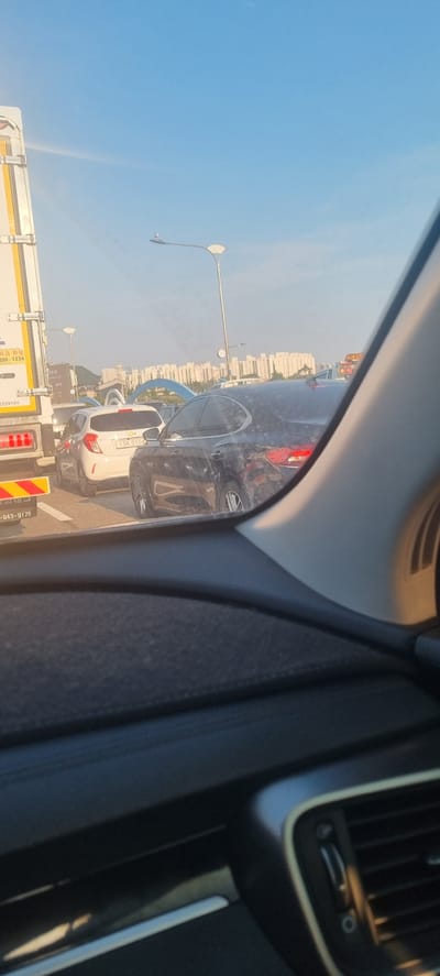 The image shows a traffic jam on a sunny day, likely in Incheon, South Korea. In the foreground, the interior of a vehicle is visible, including part of the dashboard and a side pillar. Outside the vehicle, several cars are stopped on a road. A white car with a license plate that reads "13라 0104" is prominent. To the left, a large truck with a white trailer is also stopped. In the background, buildings and streetlights can be seen, suggesting an urban or suburban setting. The sky is blue with a few wispy clouds. The overall impression is one of normal, everyday traffic congestion.