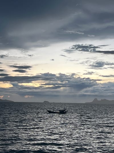 The image depicts a serene seascape with dramatic clouds and the ocean. In the water, a small, dark boat with at least two figures on board is silhouetted against the shimmering surface of the ocean. The boat appears to be moving away from the viewer. The sky is filled with a mix of dark, heavy clouds and brighter patches, suggesting either an impending storm or the aftermath of one, with a hint of sunlight breaking through. The ocean is choppy, with waves creating a textured, almost metallic sheen on its surface. In the far distance, the faint outline of land or mountains can be seen along the horizon, adding depth to the scene. The overall mood is one of tranquility and perhaps a touch of melancholy due to the dark clouds and the solitary boat. The time of day is unclear, but the light suggests it could be late afternoon or early morning. There is no visible text in the image.