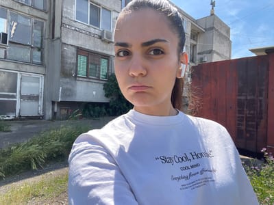 The image shows a young woman with dark hair and brown eyes, wearing a white t-shirt with text. She is standing outdoors in front of a weathered apartment building and a rusty metal fence. The building appears to be in a state of disrepair, with cracked concrete and peeling paint. The sky is blue with some wispy clouds. The overall impression is one of a somewhat neglected urban environment. There is no indication of the specific location within the city of Debelt, Bulgaria.