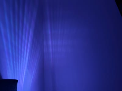 The image shows a wall illuminated by blue light, creating streaks and patterns of light and shadow. A dark object, possibly a piece of furniture, is visible in the lower left corner. The scene appears to be indoors, with the light source originating from the left side of the frame. The overall ambiance is dark and atmospheric, with the blue light casting a cool and vibrant tone across the surface. The location in Kirtorf, Germany, is not discernible from the visual information provided.