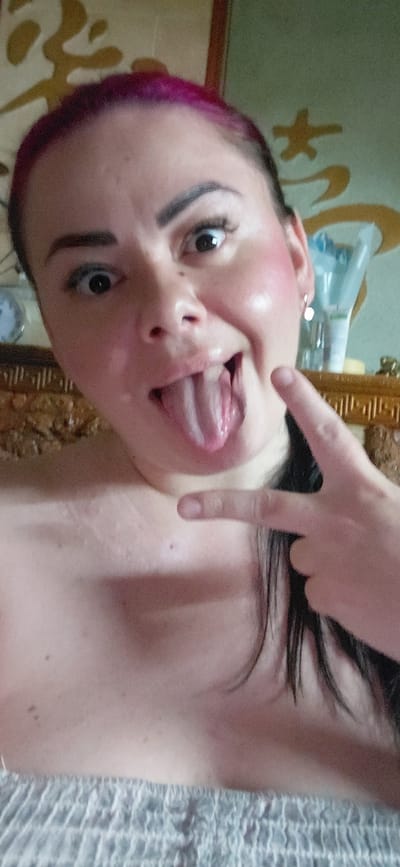 A woman with magenta hair and heavy makeup, including dark eyeliner and blush, sticks her tongue out at the camera while making a peace sign with her right hand. She is wearing a strapless garment. The background shows a wall with a decorative pattern and a shelf with various items. The image is a selfie taken from a close-up perspective.