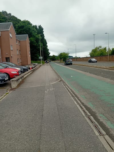The image depicts a street scene in Inverness, United Kingdom. On the left side of the frame, there are two brown apartment buildings situated against a backdrop of lush green trees and a hill. A row of parked cars is visible in front of the buildings, including a red Volvo and a dark grey Audi. A pedestrian walkway runs alongside the parked cars, bordered by a low stone wall.
The walkway transitions into a paved road with a distinct green lane, presumably for cyclists. Several cars are driving on the main road. A few pedestrians are walking on the walkway in the distance, and there are multiple streetlights lining the road. The sky is overcast, suggesting a cloudy day. A drainage grate is visible along the edge of the green lane.