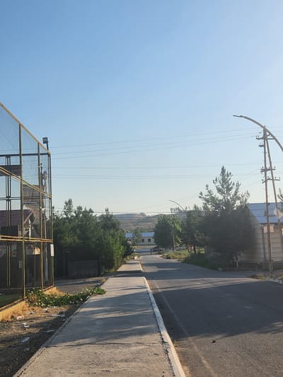 A clear, sunny day is depicted in this outdoor scene. The image shows a paved road extending into the distance, flanked by a concrete walkway on the left. On the left side of the road, a tall, chain-link fence encloses an area, with a sports facility visible behind it. Several trees line the road on both sides. In the background, there are low hills and a few buildings. The sky is a bright, cloudless blue. The scene appears to be a street or road in a somewhat rural or suburban area, possibly within the city of Xidirsho, Uzbekistan.