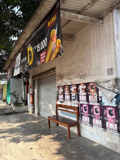 The image depicts a street scene in Kebonsari, Indonesia, characterized by a storefront with a corrugated metal roll-up door. A wooden bench sits in front of the store, adjacent to posters advertising "BASKARA SIGARET KRETEK." Above the storefront, a large black banner with yellow lettering reads "BARU" and displays a price of "Rp 15.000." The building's exterior walls are weathered and stained. To the left of the store, trees cast shadows on the concrete sidewalk. The overall environment suggests a commercial area with signs of age and wear. No people are visible in the image.