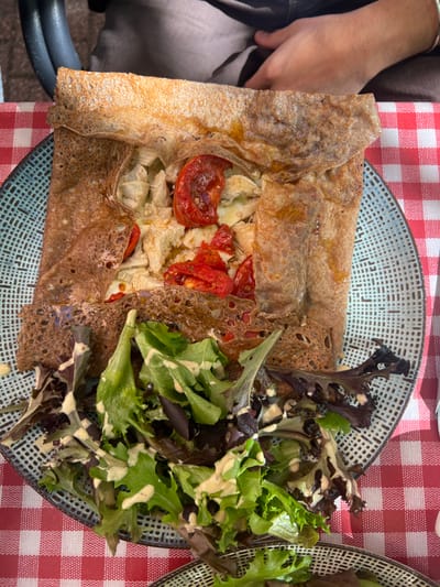 An overhead view depicts a prepared savory crêpe (galette) and side salad served on a round, textured teal-colored ceramic plate, positioned on a table covered with a red and white checkered tablecloth. The crêpe, dark brown and folded into a rectangular shape, exposes its filling of light-colored protein chunks, possibly chicken or artichoke, interspersed with bright red roasted tomato halves and a white melted substance, likely cheese. Adjacent to the crêpe, a portion of mixed green salad, comprising various lettuce leaves, is drizzled with a creamy, light-colored dressing. In the upper background, the forearm and lap of an individual, dressed in dark fabric, are partially visible, along with a dark chair back, indicating a dining setting. This scene is situated in Orléans, France.