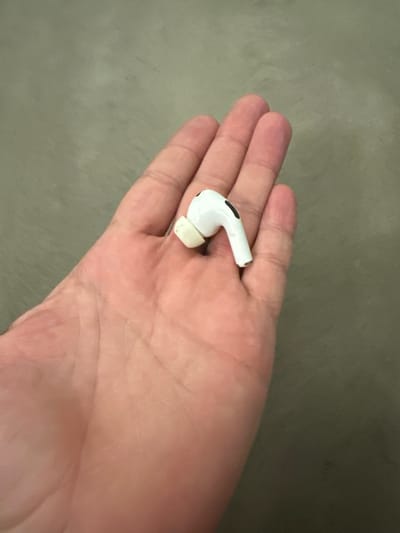 This is a close-up, top-down shot of a person's open palm, held flat and angled slightly upwards. A single white wireless earbud, likely an Apple AirPod Pro due to its distinctive white design and black accent, rests in the center of the palm. The earbud is positioned with its stem pointing away from the viewer and slightly to the right. The palm appears to be that of an adult, with visible lines and creases. The background is a muted, textured surface, likely a table or countertop, in a neutral, grayish-green hue. The lighting is even, suggesting indoor illumination, and there are no strong shadows. The image is sharp and focused on the earbud and the palm. There is no visible text in the image. The context suggests this is a personal observation, perhaps documenting a lost or found item.