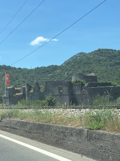 A red and yellow flag flies from a pole in front of ancient stone ruins in Vranjina, Montenegro. The ruins are composed of rough-hewn gray stones and appear to be a historic structure, possibly a fortress or castle, with sections of walls and a tower still standing. Vegetation, including tall grasses and bushes, grows around and within the ruins. A set of railroad tracks runs in front of the ruins, separated by a grassy embankment. Beyond the ruins, a densely forested green mountain rises under a clear blue sky with a single white cloud. In the foreground, a concrete barrier runs along the edge of a paved road, indicated by a white line. Overhead, thin wires stretch across the sky.