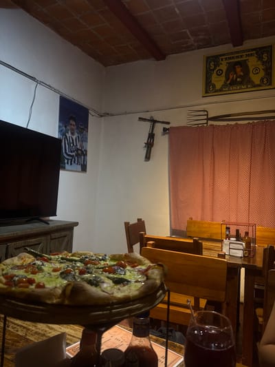 The image depicts an indoor scene, likely a restaurant or home in Chipilo de Francisco Javier Mina, Mexico, based on the decor. A large pizza is the focal point in the foreground, placed on a wooden surface next to a glass of red liquid, presumably wine. A bottle with a partially visible label is nearby, and a napkin rests against the edge of the table.
The background features various wall decorations. There's a poster resembling a dollar bill with a "Scarface" theme, a red checkered window covering, a pitchfork and a medieval-looking weapon hung on the wall, and a soccer player poster placed above a television.
The environment appears dimly lit, and the wooden furniture suggests a rustic or traditional setting. The image suggests a social gathering around food and drinks, with elements reflecting local culture and personal interests.