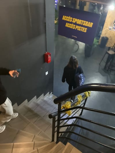 From an elevated perspective, a person walks down tiled stairs beside a black metal railing adorned with gold tinsel. A sign labeled "ACCÈS SPORTSBAR ACCÈS PISTES" stands to the left. Adjacent to it, a red box-shaped object and a small white card are positioned on the wall. Another person, holding a water bottle, is partially visible at the bottom of the frame.