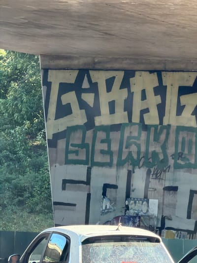 The image shows graffiti on a concrete structure, likely an overpass or bridge, with trees and foliage in the background. A white car is partially visible in the foreground. The graffiti consists of large, stylized lettering in light yellow and dark green on a grey and dark blue background. There are also some posters or paper remnants attached to the concrete wall below the graffiti. The overall scene appears to be an outdoor urban environment.