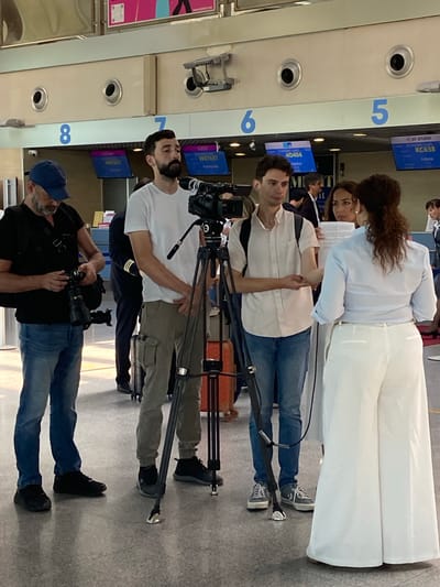 A group of individuals, including a photographer and a cameraman, are gathered in what appears to be an airport terminal in Golubovci, Montenegro. The photographer, wearing a blue baseball cap and a black vest over a black t-shirt, is holding a DSLR camera and looking down at it. Beside him, a man with a beard and a white t-shirt is standing, looking towards the front. To his right, another man in a light-colored shirt and jeans is holding a camera on a tripod, which is pointed towards a woman in a light blue shirt and white wide-leg pants. She is holding a piece of paper and appears to be speaking or being interviewed. Another woman with dark curly hair is standing behind her. In the background, there are departure boards displaying flight information, and the environment suggests an indoor public space with overhead lighting and architectural elements typical of an airport. The overall scene captures a moment of media or documentation being conducted in a travel hub.