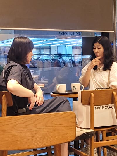 Two people are seated in wooden chairs at what appears to be a cafe or restaurant in Incheon, South Korea. The image is oriented vertically.

In the foreground, a person wearing a dark t-shirt is seated, with their arms resting on their lap. Their hands are visible, one gently touching their stomach area. A dark object, possibly a small bag or pouch, is attached to their clothing at the hip.

In the background, another person, likely a woman, is leaning their head on their hand, looking down. They are wearing a white t-shirt and have long, dark hair. A black mobile phone is held near their ear, suggesting they might be on a call or have just finished one. A white mug with the Starbucks logo is visible on a table between them, along with another white mug. A white paper bag with the text "NICE CLAU" printed in black is also on the table. Behind the people, there is a reflective surface showing a display of clothing items, possibly in a store setting. The lighting appears to be artificial and bright, with blue-toned fluorescent lights visible. The overall atmosphere seems relaxed.