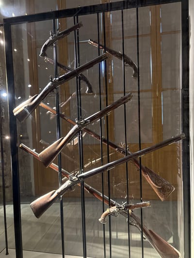 The image depicts an indoor display case containing a collection of antique firearms, specifically pistols and long guns. The weapons are arranged vertically and diagonally, appearing to be suspended by wires. The display case itself is made of metal and glass, with vertical black metal supports. The background consists of what appears to be a museum setting with wooden doors and a tiled floor.
The firearms are varied in design, featuring wooden stocks, ornate metalwork on the firing mechanisms and barrels, and some appear to have decorative engraving on the metal components. The overall lighting is dim, with some bright reflections on the glass and on the metal parts of the firearms. There are no people visible in the image.
The firearms are arranged in a manner that suggests a historical exhibition, possibly related to military history or weaponry. The style of the firearms indicates they are from an earlier era, likely pre-20th century. The context of Gatchina, Russia, suggests this may be within the Gatchina Palace, which is known for its historical collections.
