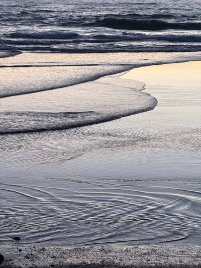 The image is a close-up, vertical shot of waves and the sandy shore at the Grande Plage in Biarritz, France. The water is dark blue with white foam crests, and the sand is wet, reflecting the light. Gentle waves create ripples and patterns on the sand as they recede. The scene is devoid of any people or animals. The overall impression is one of a calm and serene natural environment.
