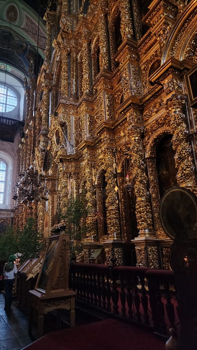 The image captures the interior of the Holy Assumption Cathedral in Smolensk, Russia. The viewpoint is looking upwards towards the highly ornate, gold-decorated ceiling. The ceiling features intricate patterns and multiple layers of decorative panels. A chandelier is visible hanging down in the center of the frame. Natural light filters in through arched windows on the lower left side, illuminating the lower portion of the interior. On the right side of the frame, there is a dark, red-covered structure, possibly a balcony or upper level section, with carved wooden details. A person is visible in the bottom right corner of the image. The overall scene conveys a sense of opulence and architectural grandeur.