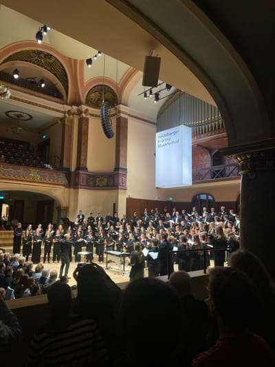 The image displays an interior view of a live musical performance occurring at the Stadthalle Heidelberg in Heidelberg, Germany, as indicated by a prominent illuminated sign displaying "Heidelberger Frühling Musikfestival." A large choir, estimated at 60-70 individuals, primarily young adults, dressed in dark formal attire, stands on tiered risers across the illuminated stage, holding sheet music. A male conductor, wearing dark clothing, stands centrally in front of the choir, facing them with outstretched arms, actively directing the performance. To the right of the conductor, a percussion instrument resembling a vibraphone or xylophone is visible with an individual standing near it. The venue features grand, ornate architecture with high arched ceilings, gold decorative accents, multiple balconies with dark railings, and a large pipe organ situated at the rear of the stage. Overhead line array speakers and stage lighting fixtures are visible. The photograph is taken from within the audience, likely from a balcony, with the backs of several audience members' heads in the foreground. One audience member in a striped shirt is actively holding up a smartphone, recording or photographing the event. The audience area is partially shadowed compared to the brightly lit stage.