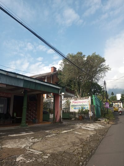 The image depicts a street scene in an unspecified location. The focus is on a building with a green roof and supports on the left. A sign for "Penginapan Syariah 145" is visible near a green gate and fence. An adjacent building has a brick chimney. A tree is positioned between the buildings. Electrical wires run across the upper portion of the frame. A yellow car and a person on a scooter are visible on the road. The road and sidewalk show signs of wear and damage. There are no visible markers indicating that this image is in Mya Sit Kaing Ywar Thit, Myanmar.