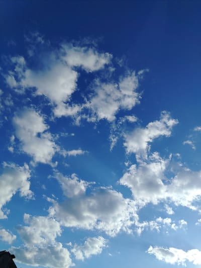 The image depicts a bright blue sky filled with scattered white clouds. Sunlight filters through the clouds, creating a dramatic effect with visible rays. In the lower left corner, the edge of a dark, angled structure, possibly a roof or a building, is visible, suggesting an outdoor scene. The overall atmosphere is serene and peaceful, indicative of a clear day. Given the location context of Zyryanskoye, Russia, this is likely a daytime scene. There is no visible text in the image.