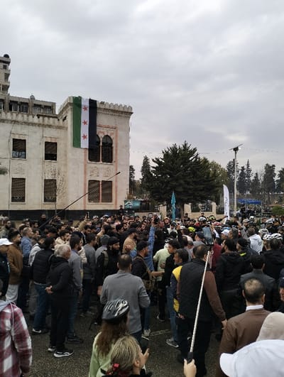 A crowd gathers in front of a building with a Syrian flag draped from its facade. People are dressed in casual clothing, some wearing helmets. Many are holding up their phones or cameras, seemingly documenting the event. The scene is overcast, with the building and surrounding trees suggesting an urban setting. The crowd's focus and the presence of the flag indicate a public gathering, possibly a demonstration or celebration.