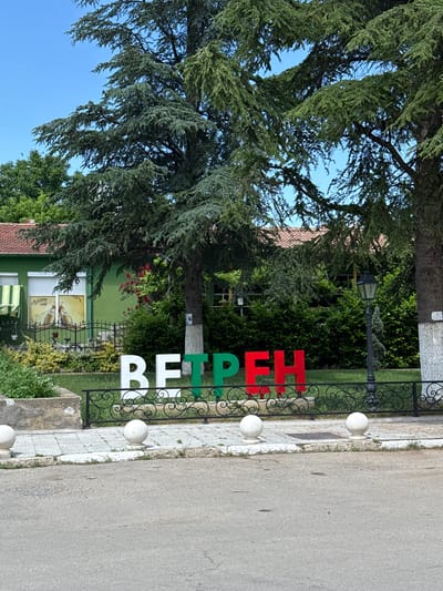 This outdoor daytime image captures a street-level view of a monument or welcome sign in Vetren, Bulgaria, indicated by the large, three-dimensional Cyrillic letters "ВЕТРЕН" (Vetren) prominently displayed in the midground. The letters are colored white, green, and red, consistent with the Bulgarian flag colors.
In the foreground, an asphalt road surface appears gray and somewhat uneven. It is bordered by a light-colored concrete sidewalk with a curb. White, spherical concrete bollards are spaced along the edge of the sidewalk. A decorative black metal fence with ornate scrollwork patterns separates the sidewalk from a landscaped grassy area.
Behind the fence, the "ВЕТРЕН" sign is positioned within a well-maintained green lawn. To the right of the sign, a black, vintage-style streetlamp with a single lamp head stands upright. Large, mature coniferous trees with dense green foliage dominate the background, some casting shadows. A two-story building with a vibrant green facade and a reddish-brown tiled roof is visible to the left. Its windows feature white roller blinds, and an awning is partially extended on the far left. One window displays a large advertisement featuring two beer bottles and the Cyrillic word "Ариана" (Ariana). The base of a tree trunk near this building is painted white, and a small, dark rectangular object is affixed to it. Further back and to the right, behind the primary trees, another building with a reddish-brown tiled roof is partially obscured by foliage. The sky above is clear and bright blue. No individuals are visible in the scene.