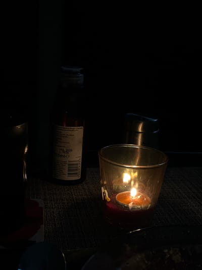The scene, located in Dorchester, United Kingdom, depicts a dimly lit tabletop environment with no visible individuals. A prominent amber-tinted glass candle holder occupies the foreground right, containing a lit tea light candle with two visible flames, resting on a red base within. To the left and slightly behind the candle holder stands a dark glass bottle with a silver screw cap, identified by its label as "Balsamic Vinegar of Modena PGI" with an expiration date of "FEB. 2028" and a volume of "215ml℮". Further in the background, to the right of the balsamic vinegar bottle, a metallic condiment shaker is partially visible. The immediate foreground displays a dark, textured surface, likely a placemat. A dark, obscured object, possibly another glass or bottle, is partially visible on the far left, and a dark, shallow dish is discernible in the bottom right.