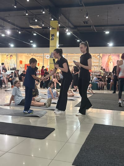 In an indoor setting, likely a mall or large retail space, a group of people are engaged in what appears to be a yoga or physical activity class. The scene is brightly lit, suggesting daytime hours. Several individuals, predominantly women, are dressed in athletic attire, including black leggings and tops. They are positioned on yoga mats, some in active poses.

A young boy in a navy blue tracksuit is a focal point, positioned on a mat and facing towards the viewer. Another young person in a light blue shirt is also on a mat, in a similar posture. The rest of the participants are spread out, with some sitting and observing, and others in various stages of stretching or exercise.

A prominent yellow banner with cartoon imagery and text is visible in the background. The text on the banner includes "moi koma". The floor is tiled and reflective, creating a mirror-like effect that duplicates some of the figures. The overall atmosphere seems focused and active, with participants demonstrating concentration and engagement in the activity. The presence of a retail background, with displays of clothing visible through glass storefronts, reinforces the idea of the event taking place in a public, commercial space. No indication of weather is discernible.