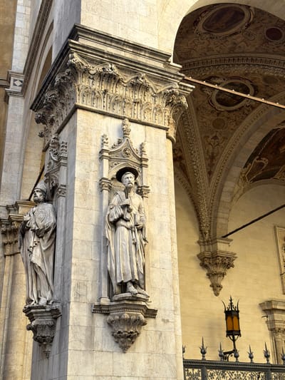 This image captures a highly ornate architectural interior, likely a cathedral or grand hall, in Siena, Italy. The perspective looks upwards, showcasing a richly decorated vaulted ceiling and elaborate wall structures adorned with sculptures.
The vaulted ceiling is covered in intricate fresco work featuring detailed patterns in muted reds, browns, and golds. Several circular medallions containing figurative paintings are visible within the ceiling design. Supporting the ceiling are massive, elaborately carved stone columns, arches, and cornices, adorned with numerous sculptural elements.
Two prominent, life-sized marble statues of robed figures are integrated into the architecture on the wall. The upper statue depicts a male figure holding what appears to be a book or scroll, standing within a decorative niche featuring a pointed, gothic-inspired canopy. Below it, another similar robed statue is partially visible. Some delicate carvings, particularly around the statues, are covered with fine protective netting.
On the far right, an antique-style, wrought-iron lantern with amber glass panels hangs from the wall, suspended by an ornate bracket. Adjacent to the lantern is a section of an elaborate metal grille with decorative, pointed finials. The overall color palette of the architecture is light stone, ranging from cream to gray, providing a striking contrast to the painted ceiling. The scene is static, highlighting the grandeur and intricate artistry of the historic building. No discernible text is visible.