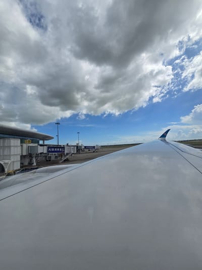 An airplane wing extends into the foreground, reflecting the sky and clouds. The wing is smooth and white, with a visible seam running along its surface. In the background, an airport terminal building is visible, with jet bridges extending out to connect with aircraft. The terminal is a modern structure with a curved roof and large glass windows. The sky is a mix of blue and white clouds, with some patches of darker clouds suggesting potential rain. The scene appears to be a tarmac at an airport, likely Shenzhen Bao'an International Airport in Bao'an District, China, given the presence of Chinese text on the jet bridges. The overall impression is one of an overcast but bright day at an airport.