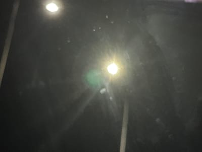 The image captures a dark, abstract scene taken at night, likely in Arad, Romania. Two prominent artificial light sources are visible: a bright, starburst-effect light in the center-left and a smaller, similar light in the bottom-right. A soft, greenish glow or lens flare appears above the main light source. The background is predominantly dark, almost black, with numerous small, blurry white specks scattered throughout, which could be dust, reflections, or atmospheric particles. Faint, elongated, light-grey shapes, possibly reflections of poles or structural elements, are discernible in the darker areas, including a distinct vertical bar on the right side of the frame. The presence of strong light sources, lens flares, and potential reflections suggests the photograph may have been taken through a window or a reflective surface. There are no people, distinct objects, or discernible activities taking place. No text is visible in the image.