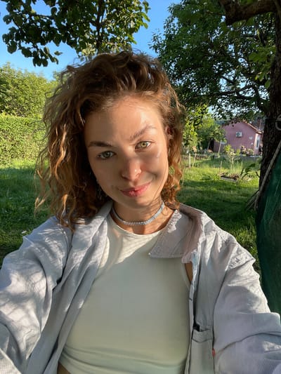 An outdoor portrait, likely a selfie, features a young woman looking directly at the camera with a gentle smile, appearing as if the image was captured with the camera rotated. Her face, framed by curly, light brown hair, occupies the foreground on the left, revealing striking green eyes and a natural sheen on her skin, possibly from sunlight. She wears a white tank top beneath an open, light grey/blue plaid patterned button-up shirt, accessorized with a sparkly choker necklace.

The background depicts a vibrant garden or backyard scene in Danilovgrad, Montenegro, under a clear blue, sunny sky. Large trees with dense green foliage dominate the upper and left parts of the frame, casting dappled shadows. In the mid-ground, a distant house with pinkish-terracotta walls and an visible air conditioning unit is nestled among more lush greenery. A green netting or tarp is partially visible in the upper center. The bright daylight and verdant surroundings suggest a pleasant, natural environment. No visible text is present in the image.