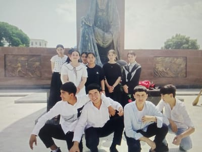 This is a photograph of a group of nine young people, likely students, posing in front of a monument. The monument appears to be a large, draped statue of a person, set against a background of a stone wall with relief carvings. Behind the group, a building with visible windows can be seen. The people are arranged in two rows: five are in the front, crouching or kneeling, and four are standing behind them. The individuals in the front row are all male, dressed in casual shirts and dark pants, with two of them interacting with each other. The individuals in the back row are all female, dressed in various tops and dark skirts or pants. The environment suggests an outdoor public space, possibly a memorial park or a historical site. The overall lighting indicates it is daytime, with a bright, overcast sky. The location is identified as Karaganda, Uzbekistan, although Karaganda is located in Kazakhstan, not Uzbekistan.