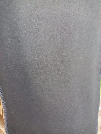 The image shows a close-up view of a black fabric with a ribbed texture. The fabric appears to be synthetic and is likely part of an article of clothing or a textile item. There are some small white specks, possibly lint or dust, visible on the surface of the fabric. The edge of the fabric on the left side shows a stitched seam. The background is mostly out of focus, but hints of green suggest an outdoor environment.
There is no information or visual cues in the image to suggest a specific location in Zinna, Nigeria. The image focuses solely on the texture and detail of the black fabric.