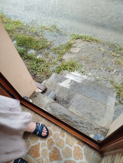 The image shows a view from an open doorway, looking out onto a set of stone steps that are wet and appear to be experiencing flooding from the rain. A person's legs and feet are visible in the foreground, wearing dark blue sandals and light-colored pants or a skirt. One foot is on a tiled floor inside the doorway, and the other is partially visible near the steps.
The scene is outdoors, with grass and some debris visible on the ground beside the steps. The ground is saturated with water, and water is actively flowing down the steps. The weather appears to be rainy or has recently rained, contributing to the flooded condition. The doorway has wooden framing, suggesting it's an entrance to a house or building. The overall lighting suggests it is daytime, but the overcast weather due to rain likely makes it appear somewhat dim. The setting appears to be a residential outdoor area. There is no visible text in the image.