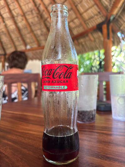 A partially consumed, returnable glass Coca-Cola Zero Azúcar bottle stands prominently on a dark, polished wooden table, covered in condensation, indicating it was recently cold. The bottle contains a dark brown liquid, approximately one-quarter of its capacity. To the left, a clear drinking glass is partially visible. To the right, two additional clear cups are present; one contains a dark granular substance at its base, and the other appears mostly empty. In the blurred background, the back of an individual with short, dark hair is visible, seated at a wooden table beneath a thatched roof structure (palapa) supported by dark wooden beams. Lush green foliage is discernible in the far background. The scene is located in an outdoor dining area in Cabo Velas, Costa Rica.