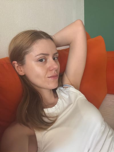 The image shows a young woman with light brown hair lying on orange pillows. She is wearing a white tank top. The background consists of a textured wall, part of which is painted green. The woman is looking at the camera and has her arm behind her head. There are no discernible landmarks or elements that would place this image in the city of Helmond, Netherlands. The scene appears to be an indoor setting, possibly a living room or bedroom.