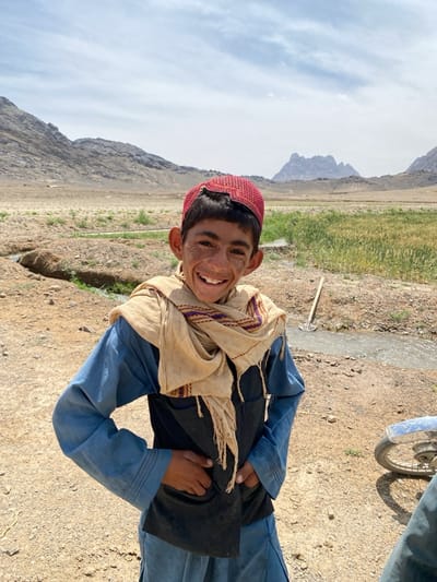 A portrait-style photograph features a young boy, likely in his early teens, standing outdoors and smiling broadly at the camera. He has dark hair, a wide, joyful grin, and prominent ears, with some visible dirt or freckles on his face. He wears a red, intricately embroidered cap, a light blue long-sleeved tunic (likely part of a shalwar kameez), and a dark blue or black sleeveless vest over it. A light beige scarf with purple and red patterned borders and fringes is wrapped around his neck and shoulders. His hands are placed casually on his hips.
The boy stands on a light brown dirt path or ground. The background reveals a vast, rugged mountainous landscape under a bright, partly cloudy sky. Patches of green fields or crops are visible on both sides of the boy, suggesting agricultural activity. A narrow water channel or stream runs through the mid-ground. In the distance, sharper peaks of mountains can be seen. On the far right, part of a motorcycle, including its front wheel and fender, is visible. A long-handled tool, resembling a shovel or hoe, leans near the stream on the right. The bright, clear daylight suggests it is midday or early afternoon. The boy's expression conveys happiness and friendliness. There is no visible text in the image. The scene depicts a rural, arid region, consistent with the location context of Ashoqah, Afghanistan.