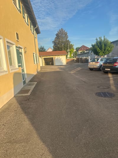 The image captures a sunlit scene in Mainburg, Germany, likely in the late afternoon or early evening. In the foreground, the yellow facade of a building is illuminated, casting a long shadow across the asphalt-paved area. The building features several windows with green shutters. Adjacent to the building, a rectangular patch of concrete with two manhole covers is visible.
Further into the scene, a low-slung building with a red-tiled roof houses a garage with two white doors, one of which is open. Inside the garage, various items are stored, including a bicycle. Beyond the garage, a metal fence with a diamond pattern encloses an area.
Several cars are parked in the paved area. A silver hatchback with the license plate "KEH C 306" is parked facing away from the camera. To its right, a dark-colored hatchback with the license plate "MAI AC 777" is also parked.
The background reveals a residential area with other buildings, trees, and a bright blue sky with scattered white clouds. The overall impression is one of a quiet, residential street bathed in the warm glow of the setting sun.