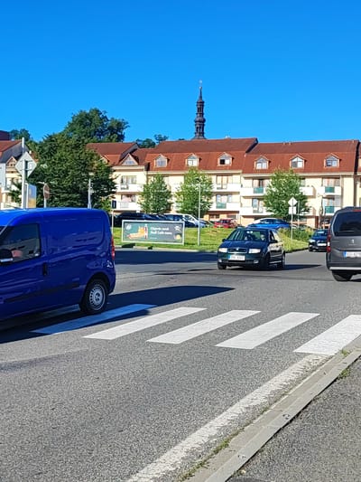 A blue van is parked in front of a pedestrian crossing on a sunny day. The van is in the foreground, on the left side of the image. The pedestrian crossing is marked with white stripes on the asphalt road. In the background, there are several cars, including a dark green Skoda driving on the road, and a grey van on the right side of the image. Behind the cars, there is a large, light-colored building with a red tile roof and many windows. A tall, dark church spire can be seen rising above the building and trees. The sky is clear and blue, indicating a sunny day. A billboard advertising "Objednejte nová Shell Café menu" (Order the new Shell Café menu) is visible to the right of the blue van, showing a hot dog and coffee. The scene appears to be a street in a town or city, likely during daytime given the bright sunlight. The overall impression is one of everyday traffic and urban scenery.