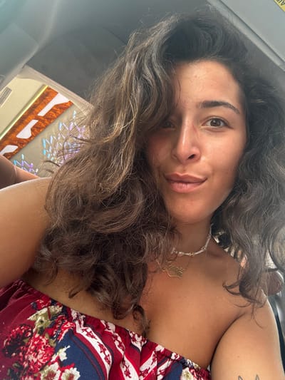 The image is a selfie taken by a young woman with dark, wavy hair. She is wearing a strapless red top with a floral pattern. She has a necklace with a gold pendant around her neck. Her skin is tanned, and she has light makeup on. She is smiling slightly and looking at the camera. The background appears to be inside a vehicle, with a glimpse of what looks like a digital display or advertisement featuring bright colors and geometric patterns. The lighting suggests it is daytime. There is no discernible information in the image that places the location in Kompung Soeng, Cambodia.