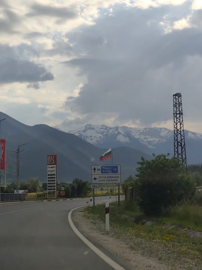 The image captures a roadside scene near Dobrinishte, Bulgaria, identifiable by the road signs, a Lukoil gas station, and the Bulgarian flag. The road curves slightly, leading past a grassy area dotted with yellow and white wildflowers. A sign indicating "Razlog 13" and "Centre Dobrinishte" is visible. In the background, mountains with snow-capped peaks are prominent under a cloudy sky. A latticed metal tower stands on the right side of the frame, adding to the industrial elements in the landscape. The environment includes a mix of natural and man-made features typical of a roadside in a rural or semi-rural location.