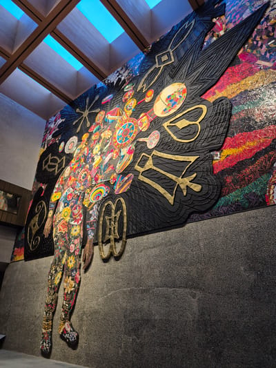 This is a photograph of a large, colorful mural with a three-dimensional effect mounted on a dark gray textured wall, likely in an indoor public space such as a gallery or atrium. The mural depicts a stylized, elongated figure composed of various abstract shapes, floral patterns, and circular motifs in vibrant colors like red, yellow, blue, and pink. The figure's head is a cluster of these colorful elements, and its body appears to be a cascading arrangement of floral designs. Emerging from the upper part of the figure are large, black, wing-like shapes with gold-colored symbolic inscriptions. The mural has a sense of depth due to the layered elements and textures. Above the mural, a ceiling with wooden beams and rectangular skylights reveals a bright blue sky, suggesting the space is open to natural light. The lower portion of the image shows the textured gray wall and a hint of a wooden baseboard. The overall impression is one of a dynamic and intricate art installation. This artwork is located in Princeton, New Jersey, United States of America, specifically within the Art Museum at Princeton University.