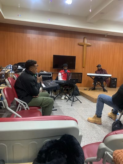 The scene is located in Shenyang City, China, within an indoor space consistent with a church or multipurpose hall. The room features light brown wooden paneling on the back wall and a light gray tiled floor. A prominent wooden Latin cross is centrally mounted on the back wall. The ceiling is off-white with recessed lighting, from which several iridescent, decorative ornaments are suspended.

Approximately five individuals, all appearing to be male, are present. In the foreground left, a man in a black puffer jacket and olive green cargo pants is seated in a red and off-white chair, holding a microphone in his left hand and resting his right hand on a small black electronic keyboard or MIDI controller on his lap. To his left, partially obscured, another man in a plaid shirt is seated behind a drum set, with a bass guitar nearby. In the midground, a man wearing a red and white jacket and a black beanie is seated behind a black digital keyboard on a stand, positioned in front of a music stand. A large flat-screen television is mounted on the wall above this individual. To the right, on a low wooden platform, a man in a dark cap and jacket is seated behind a white digital keyboard on a stand, with a dark speaker positioned to his right. In the foreground right, the lower body of a fifth person is visible, wearing blue jeans and light brown work boots, seated in a similar red and off-white chair.

The individuals are engaged in a musical activity, likely a rehearsal or performance, with three actively playing instruments or using a microphone. The arrangement of people and instruments suggests a band or worship team setting.
