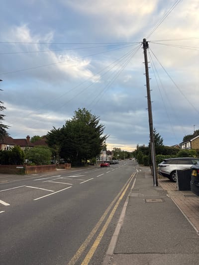 The image depicts a residential street scene in Chelmsford, United Kingdom, at dusk. A tall wooden utility pole with numerous wires extending from it dominates the right side of the frame. The street is asphalt, with double yellow lines marking the center and white road markings indicating a pedestrian crossing ahead. Several cars are parked along the side of the road, including a silver SUV. On the left side of the street, there are houses with brick walls and gardens. The sky is cloudy, with hints of sunset colors in the distance. A street sign labeled "THE CEDARS" is visible on the left.