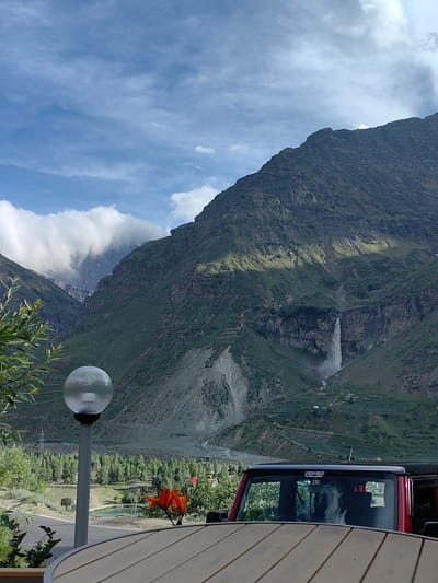This image captures a scenic mountain landscape with a waterfall cascading down a steep slope in the distance. In the foreground, a red vehicle is partially visible, with its roof and windshield in view. A wooden table is also in the foreground, suggesting an outdoor seating area. A lamppost with a globe-shaped light fixture stands to the left of the frame, and some green foliage is present in the lower left corner. The background is dominated by lush green mountains, with a river flowing through the valley. The sky is partly cloudy, with patches of blue visible. The location is identified as Yamling, India.