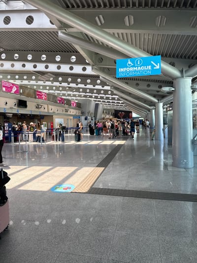 The image depicts the interior of an airport terminal in Golubovci, Montenegro. The scene is brightly lit by natural light streaming in from large windows, creating distinct shadows on the polished floor. The architecture features a modern industrial design with exposed metal beams and a corrugated metal ceiling.

In the foreground, a light pink suitcase is partially visible on the left. The floor is tiled with a gray, speckled granite pattern. A tactile paving strip runs horizontally across the mid-ground, indicating a pathway or hazard warning. A circular floor decal with colorful cartoon figures, possibly a social distancing reminder, is visible on the floor in the lower-middle section.

The middle ground shows a queue of people, mostly standing with luggage, waiting at check-in counters. These counters are numbered from 2 to 5. Various signs and advertisements are displayed above the counters, some featuring pink branding. People are dressed casually, and most have carry-on luggage.

In the background, more people are visible, some moving through the terminal, others standing. The architecture continues with large, silver cylindrical columns supporting the overhead structure. A bright blue sign with an accessibility icon, an information icon, and the words "INFORMACIJE INFORMATION" with a white arrow pointing right is attached to a column, indicating directions. Further in the background, several glass-enclosed areas or doorways are visible, suggesting other sections of the airport. The overall impression is one of a busy, functional airport environment.
