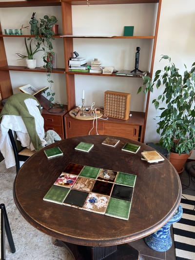 The image depicts an indoor scene, likely a living room or dining area, featuring a round wooden table with several square tiles arranged on its surface. The tiles vary in color and pattern, with some being a solid dark green, others a lighter green, and a few with intricate colorful designs. Surrounding the table, a striped rug is visible on the floor. To the right of the table, a large potted plant with green leaves stands in a terracotta pot. In the background, a wooden shelving unit is filled with books, decorative items, and a small green square object. A white ceramic candle holder with a lit candle is also present on a tray on a wooden cabinet to the right. A chair draped with a green sweater and white fabric is positioned to the left of the table. The overall impression is one of a cozy, decorated domestic space. The location in Hämeenlinna, Finland is not discernible from the visual elements present in the image.