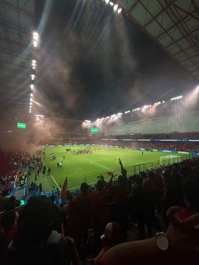 The image depicts a soccer game inside a stadium, likely in Toluca, Mexico based on the stadium design and the team colors. The stadium is densely packed with spectators, many of whom are wearing red, which might be the team color. There is smoke or pyrotechnics being used, obscuring parts of the stadium lights and creating a dramatic effect. The field is green, and players and officials are visible. The camera angle is from an elevated position within the stands, possibly looking down at the field. A person wearing a wristwatch is partially visible in the foreground, indicating that the picture was taken by someone at the event. Banners with sponsors logos (aramco,Corona) are visible along the side of the field. The event seems lively and festive.